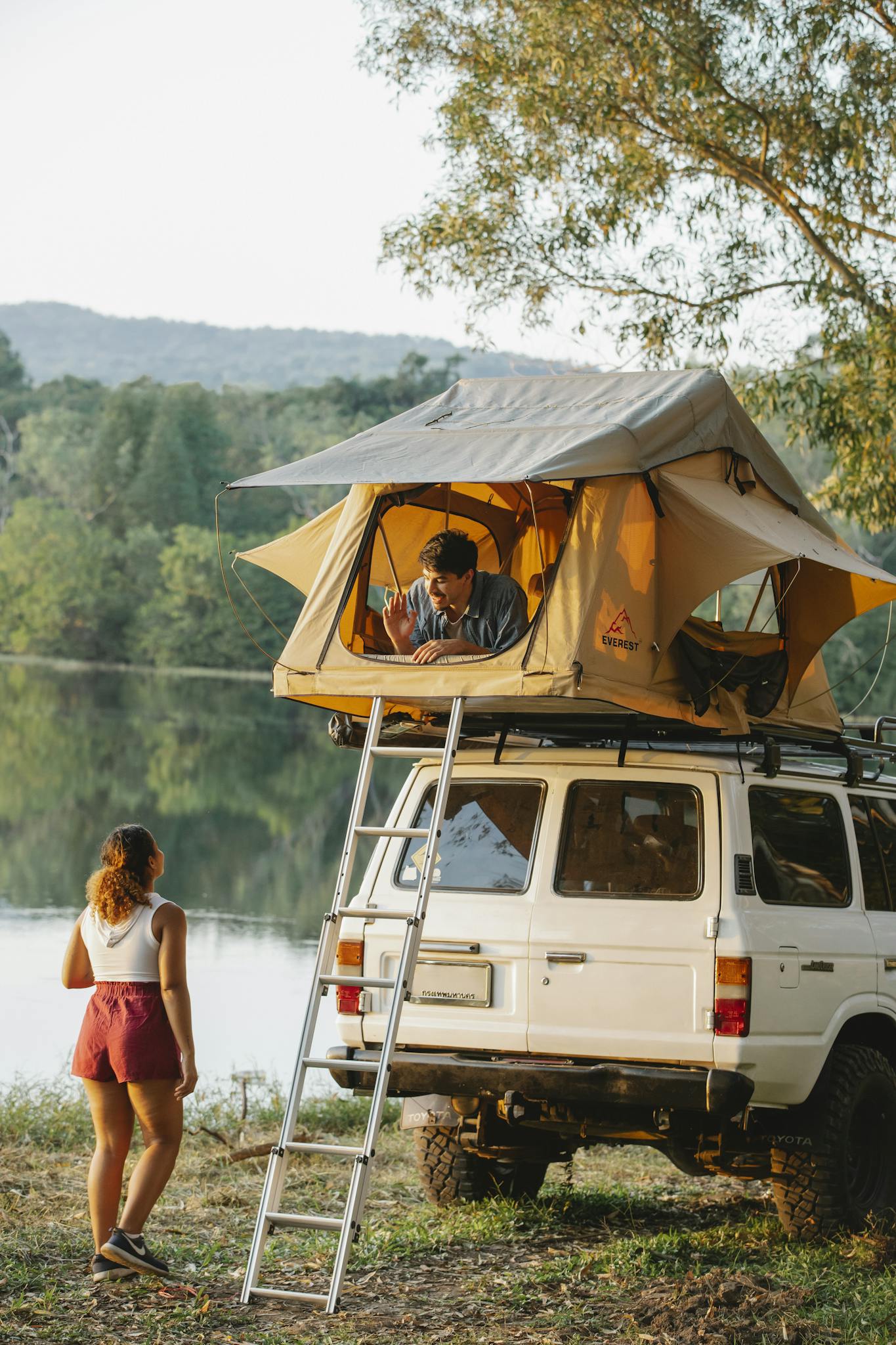 Back view of unrecognizable young female traveler standing on lake shore and talking to boyfriend resting in tent placed on SUV car roof